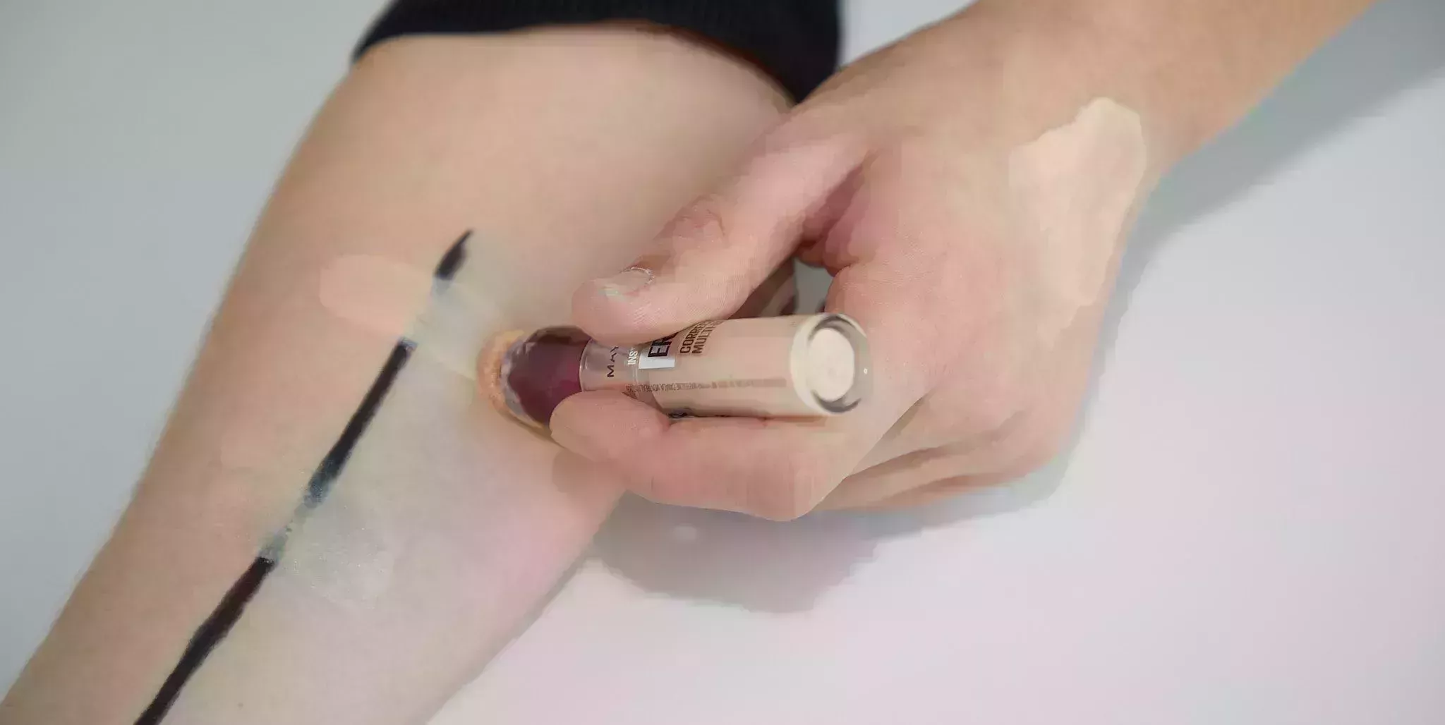 a medium coverage and sheer coverage concealer being swatched on top of an arm with a stripe of eyeliner on it