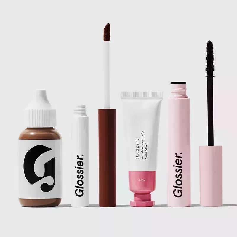 A photo of Glossier products, including the Perfecting Skin Tint, Skywash liquid eyeshadows, Cloud Paint cream blush, and Lash Slick mascara on a gray background