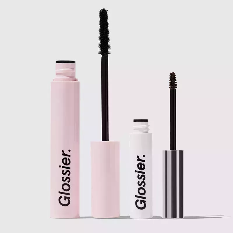 A photo of the Glossier Lash Slick mascara and Boy Brow eyebrow gel on a gray background