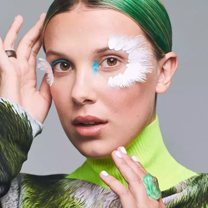  Portrait of Millie Bobby Brown for Allure. She wears a white arm cuff made of paper that obscures half her face. She’s also wearing silver eye makeup and her hair slicked back.